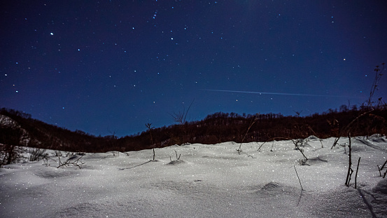 Nighttime scenery in natural environment with mountain and village lights in the background during winter season and white snow covering the ground with light moon and starry sky.