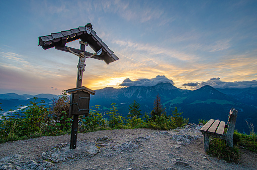 Kreuz mit Jesusfigur auf dem Gipfel des Grünstein im Nationalpark Berchtesgaden vor dramatischem Himmel kurz vor Sonnenaufgang