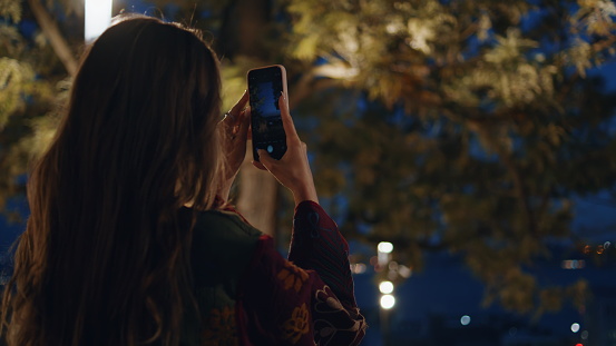 Girl photographing night park on smartphone close up. Attractive young woman taking picture green town nature standing at twilight alone. Relaxed lady using mobile phone recording video dark city.