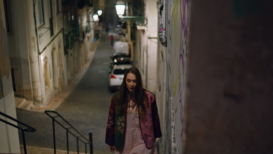 Woman traveler going up stairs night city street. Carefree happy girl walking upstairs exploring empty town lane alone. Gorgeous young lady tourist strolling late evening wearing vintage jacket.