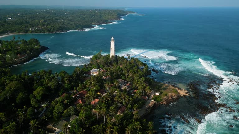 Aerial drone view of historical Dutch lighthouse on tropical island surrounded by greenery, with powerful ocean waves, clear blue skies, tourists explore shore, nature raw vintage maritime landmark.