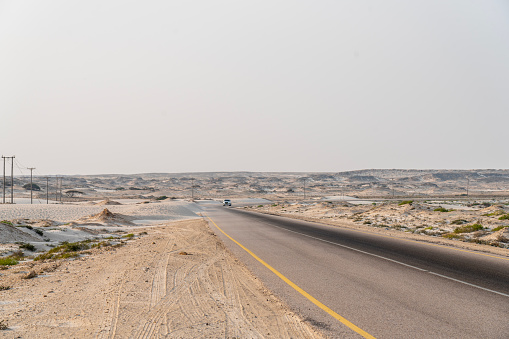 Empty diminishing asphalt road, on both sides endless white salty plain with electric poles against blue sky. In distance Sugar dunes at Al Khaluf, Oman