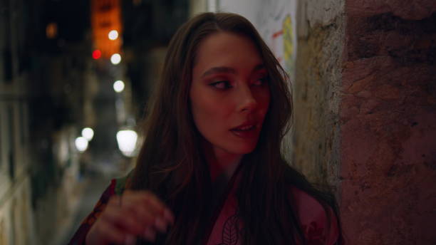 Gorgeous model standing urban street late evening looking camera confidently close up. Portrait of attractive glamorous woman leaning at city building wall in front night city lights. Town nightlife.