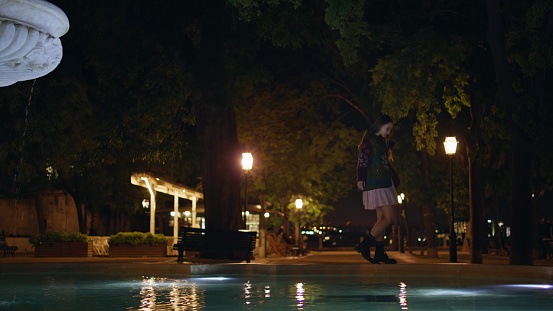 Girl walking on fountain edge calm summer night. Cute trendy woman balancing over water reflecting street lights. Carefree young lady relaxing in dark evening town square alone enjoying nightlife.