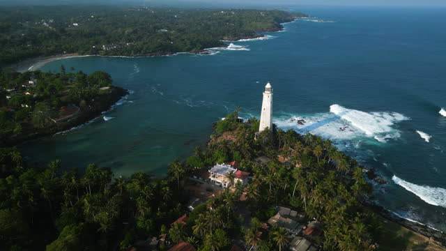 Aerial drone of Dondra lighthouse surrounded by palm trees in Sri Lanka. Waves crash against rocks, boats navigate nearby waters, serene beauty attracts travelers seeking picturesque coastlines.