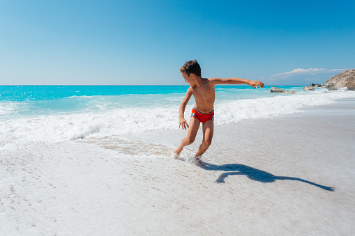 Teenage boy enjoys summer vacation running and jumping on the waves at a beautiful beach on the Greek Lefkada island