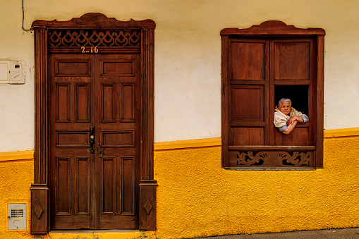 Jerico, Colombia - January 14, 2023: Elderly woman peeks out of the window of her colorful house