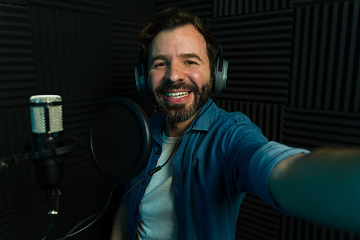 Cheerful man with headphones taking a selfie while recording in a soundproof podcast studio