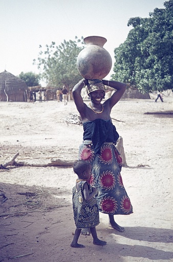 Ghana (exact location not known), West Africa, 1973. Woman with clay jug on her head and child in front of a Ghanaian village. Furthermore: travelers.