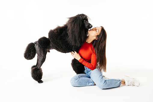 A woman shares a loving kiss with her poodle, both enjoying a playful moment on a white background.