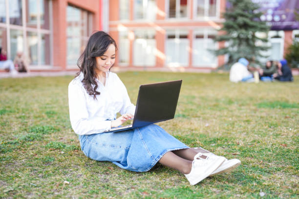 istock Young female student sitting on the grass at university campus studying on laptop 2082004699
