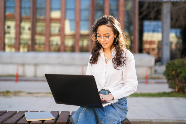istock Asian young female student working with laptop sitting on bench at university campus 2082006273