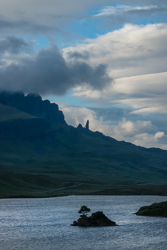 SILHOUETTE: Tiny islet in Loch Fada and great Old Man Of Storr shrouded in clouds. Dramatic mountainous landscape with majestic peaks towering above wavy sea water on picturesque Scottish Isle of Skye