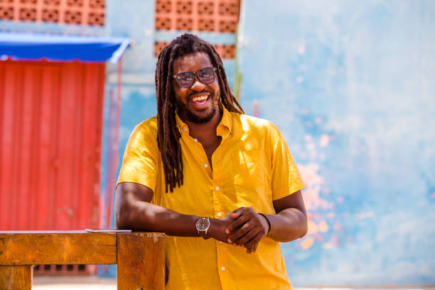 stylish man on a walk through the city smiles looking at the camera. A black man with afro hair is leaning against the railing, wearing a yellow shirt, sunglasses and a watch.