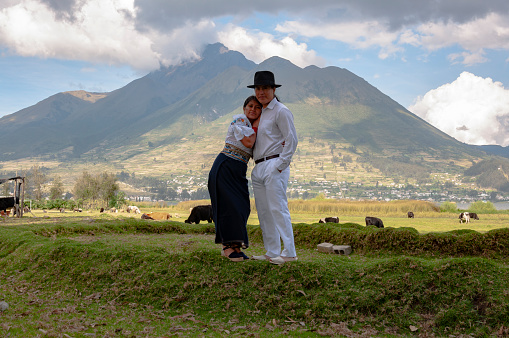 A couple dressed in traditional Andean clothing stands closely together in a rural highland field in Ecuador, with the imposing backdrop of a volcanic mountain. The woman, holding a small bouquet, leans against the man, who is dressed in a white suit and a black hat, indicative of local cultural attire.