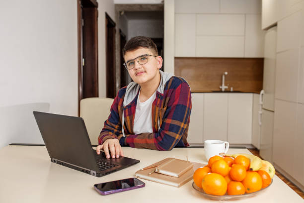 Teen Boy with Glasses Working on Laptop at Kitchen Table. Gen Z.