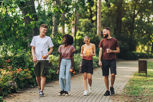 Full length of multiracial friends taking a walk in city park with beverages in their hands and spending quality time together. An interracial group of trendy hipster friends going for a walk in park.