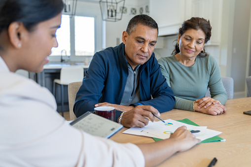 A couple sit at the kitchen table with a life insurance agent as they review their coverage and plan for the future.  They are both dressed casually  and appear content.