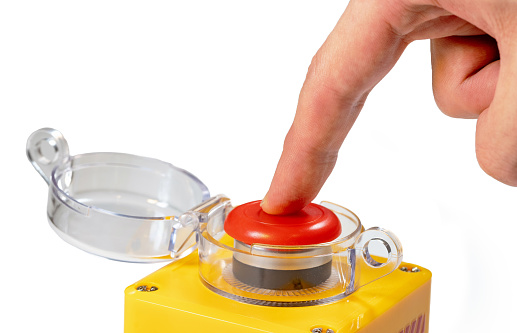 Finger pressing down on a large red yellow button with a clear plastic lid isolated on white background man pressing an emergency industrial machinery stop button, closeup, one person, safety protocol