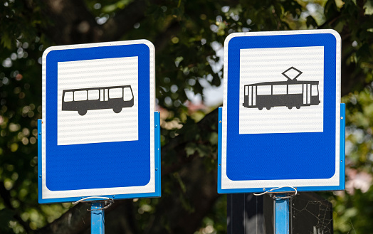Blue bus and tram stop signs side by side, object closeup, against a blurred tree background, daylight, nobody. City transportation commute, means of eco transport abstract concept, two signs together