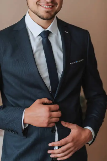 Photo of Confident elegant handsome guy standing at home and buttoning his jacket looking at the camera in a nice dark blue suit