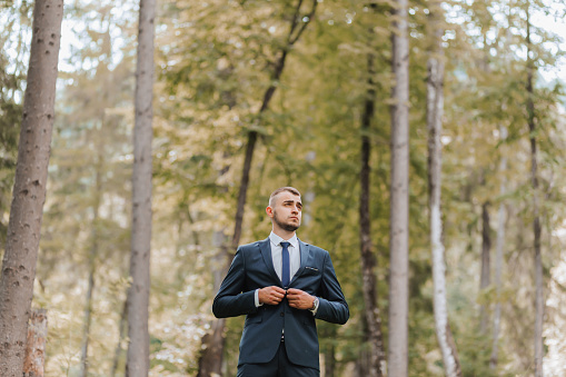 A young man in a classic suit against the background of tall trees in a forest or park. The groom fastens his jacket