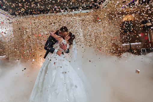 Gorgeous bride and stylish groom dancing under golden confetti at wedding reception. Happy wedding couple performing first dance in restaurant. Romantic moments