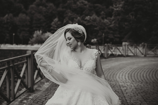 Wedding portrait. The bride in a white dress on the bridge with a flowing veil, the groom is standing behind her. Sincere smile. Wind and veil. Diadem. Black and white photo