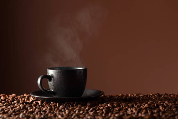 A warm brown bowl filled with freshly roasted coffee beans emits steam beside a steaming cup of brewed coffee against a black backdrop