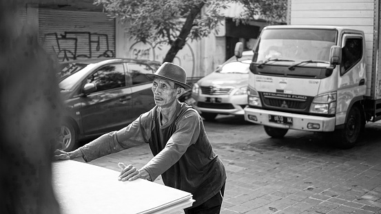 a middle-aged man in a bowl hat is working, pushing his wares, on Jalan Braga, Bandung, West Java, Indonesia.