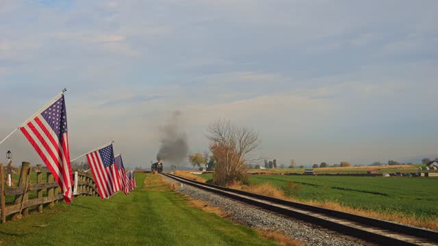 View of a Single Rail Road Track, With a Steam Passenger Train Approaching, With a Fence with America Flag on it, Gently Waving in the Wind on a Sunny Autumn Day