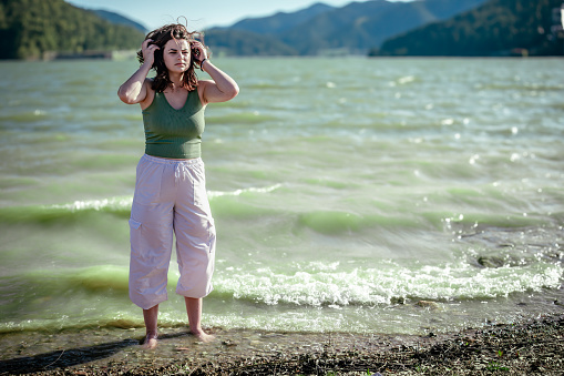 woman in white pants and green crop top stands on shore of choppy lake with mountains in the background