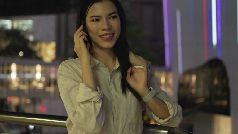Businesswoman standing on the sky train station talking, speaking, chatting on brand name smart phone while waiting for the train after work overtime. Bangkok, Thailand.