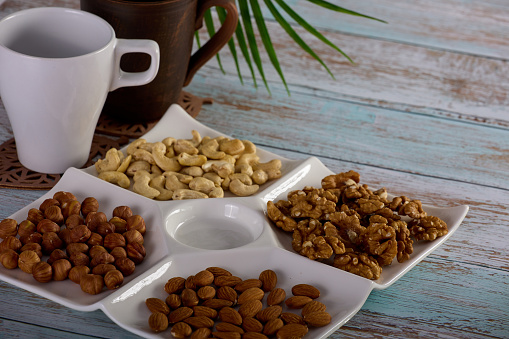 Dried nuts of various types are beautifully placed on a square saucer standing on a wooden table. Assorted nuts are laid out on a square-shaped plate along with empty glasses for hot drinks.