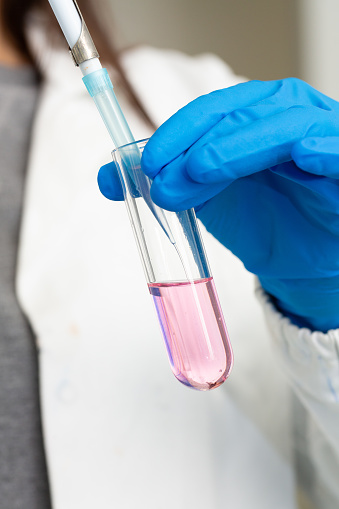 A scientist's gloved hands pipetting a pink chemical solution into a clear test tube in a lab setting