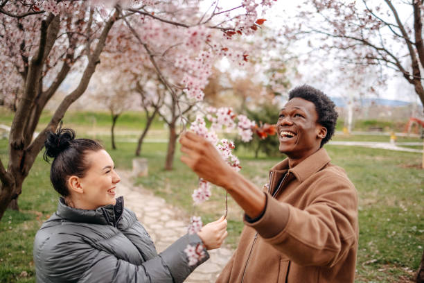 multiracial couple enjoying spring walking in city park full with tree flowers - friendship park flower outdoors stock-fotos und bilder