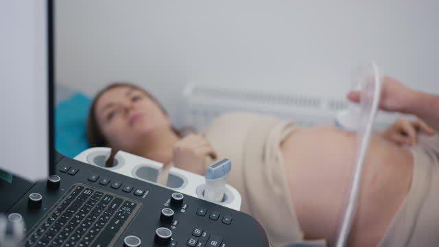 Close-up. An unrecognizable male ultrasound specialist turns the monitor screen towards a pregnant woman, who is lying on her back on a medical couch, during an ultrasound procedure.