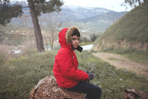 Adorable teenager boy traveler in red parka, holding a thermos mug with hot drink, looking at the camera, sitting on a log in the forest, relaxing while trekking in mountains on the early spring day