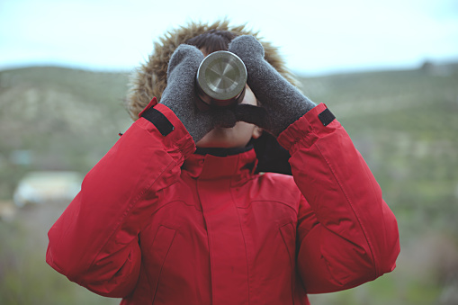 Teenager boy in red parka with fluffy further hood, drinking from steel travel thermos mug against mountains background. Child adventurer drinks hot tea in early spring nature, cold weather outside