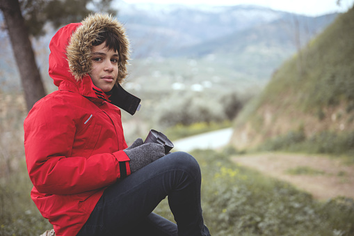 Teen boy holding a thermos mug, taking a break while trekking in mountains. Handsome teenage boy in warm red parka relaxing, sitting on a log, discovering beautiful early spring nature. Active travel.