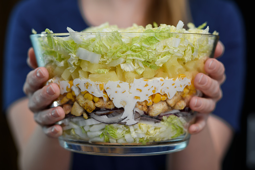 Woman holds in her hands a glass bowl with layered party salad with chicken and pineapple
