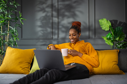 Portrait of beautiful young African woman enjoying cup of coffee while sitting on sofa and using laptop at home