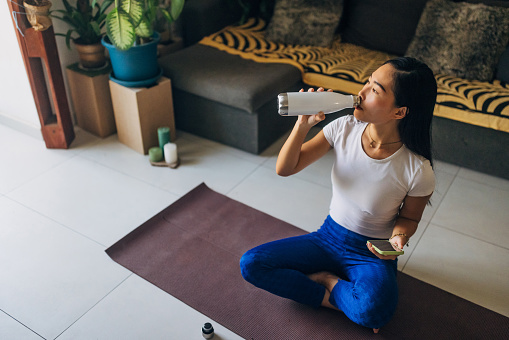 Taiwanese woman sitting on exercise mat in living room, using mobile phone and drinking water from stainless steel water bottle