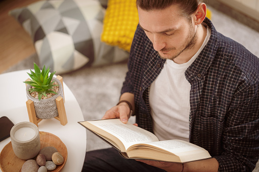 High angle view of a young Caucasian man sitting in the living room reading a book