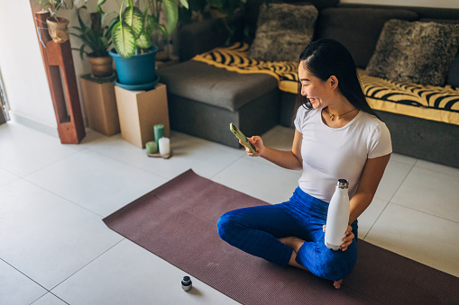 Cheerful woman taking a break from meditation sitting on an exercise mat, using a mobile phone and drinking water from a stainless steel bottle