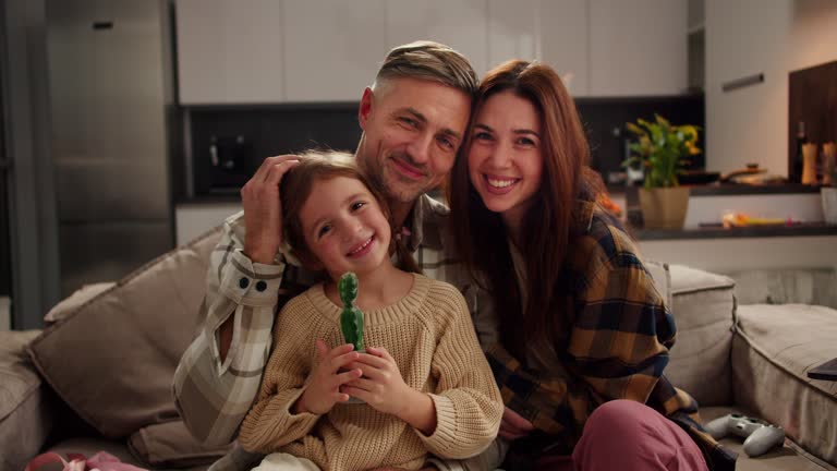 Portrait of a happy family a little brown-haired girl in a cream sweater holds a cactus toy in her hands and sits with her parents a man with gray hair and a brunette woman in a plaid shirt on the sofa in a modern studio in an apartment in the evening