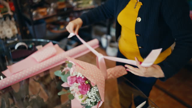 Smiling Mature Female Florist Tying Pink Ribbon on Beautiful Bouquet of Flowers in Shop