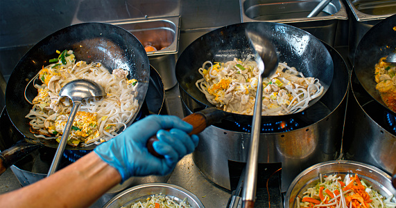 Male chef in an Asian restaurant tending to three dishes in woks on the gas stoves.