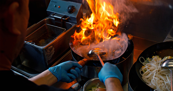 Male chef in an Asian restaurant mixing the dish in the wok as it catches fire for a short amount of time.
