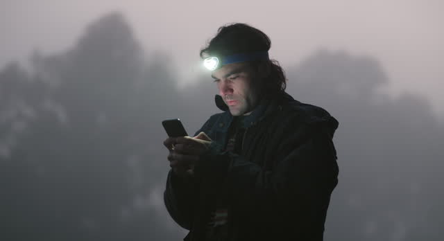 Man, forest and phone in night, texting and torch on head for vision, thinking or lost with communication at camp. Guy, smartphone and lamp for light, search and map app for hiking adventure in woods